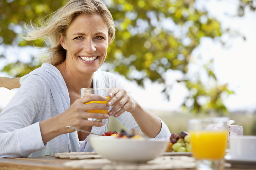 Smiling mature woman with orange juice at breakfast table outdoors - summertime hygge