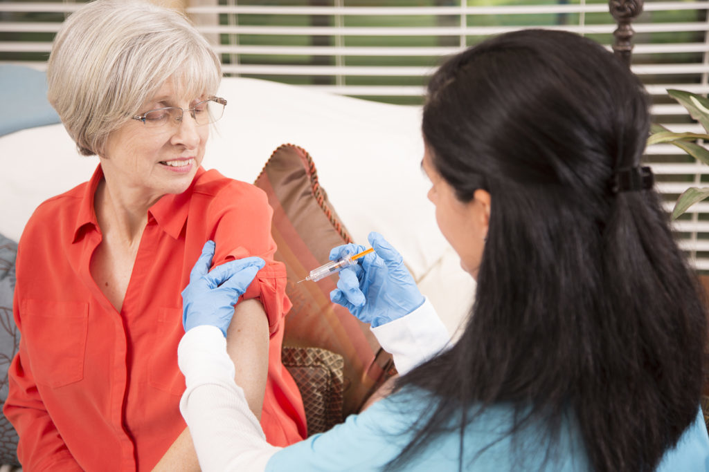 Senior adult, female patient receives vaccine or medicine from her Latin descent, home healthcare nurse in nursing home or home setting. Nurse wears gloves and holds syringe.