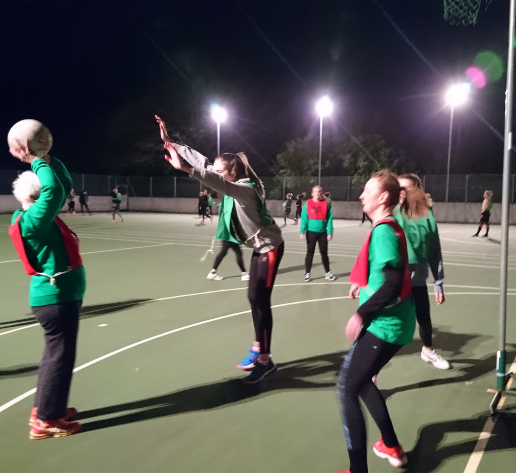 Elderly lady holding netball above her head as she aims to throw it into the basket, other players defending