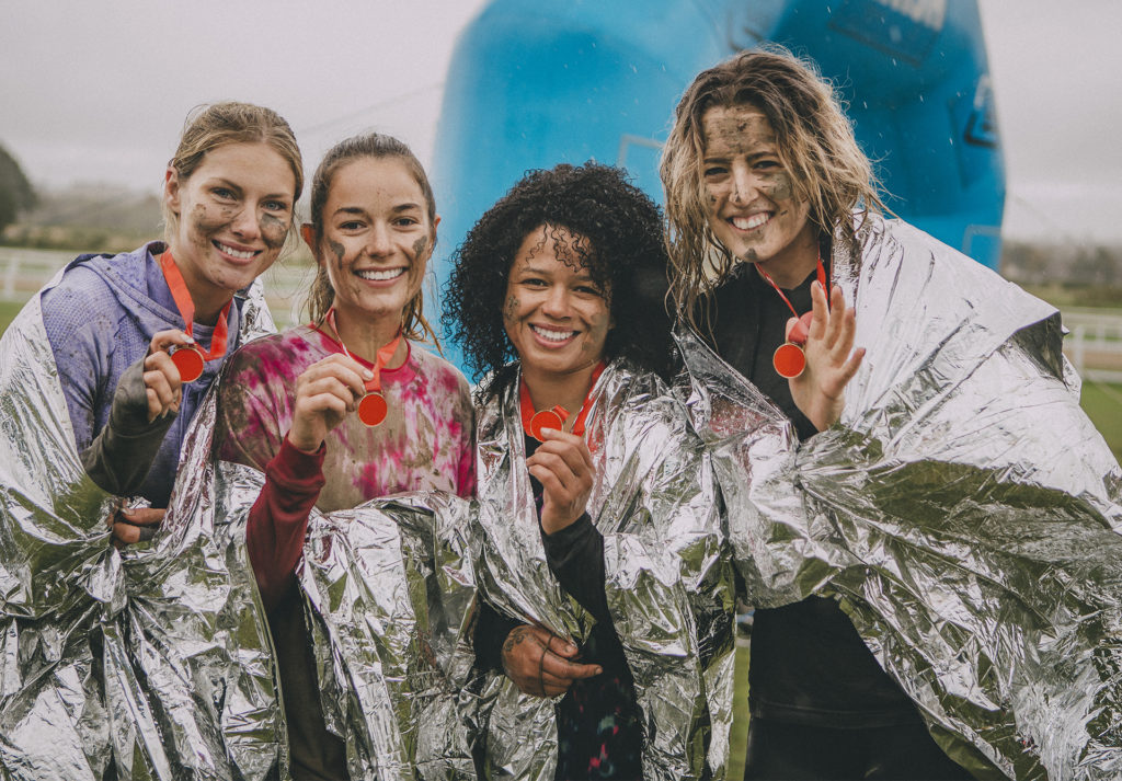 Four women are posing for the camera in space tech foil blankets with the medals they have won from completing a charity obstacle course.