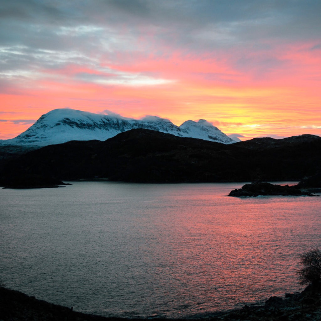 Hills, some snow-covered, at sunset, pink light reflected in sea