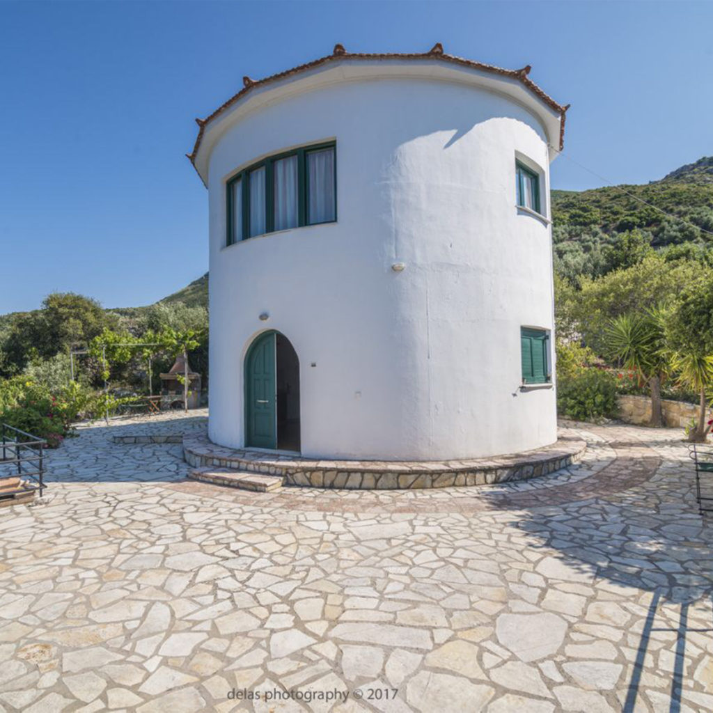 White painted circular building, Greek windmill converted into holiday accommodation, hills behind, stone terrace in front