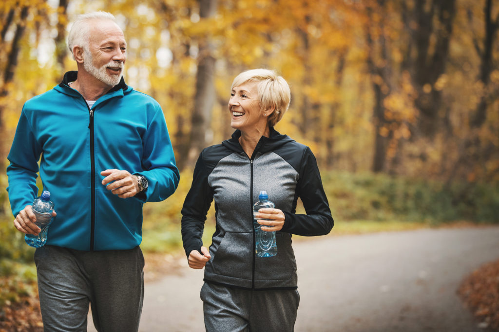Closeup front view of a senior couple jogging in a forest and having fun. They are running on a winding forest road, laughing and doing their healthy routine. Both holding water bottle.Trees in background have turned orange and yellow and there's a lot of leaves on the side of the road.
