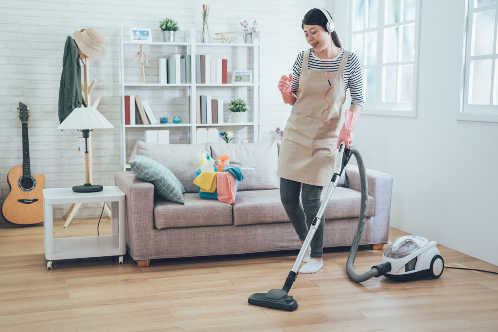 Smiling excited young housewife havig fun while cleaning floor with vacuum cleaner. Happy woman doing housework at home enjoy music wearing earphones. lady in apron singing dancing house chores