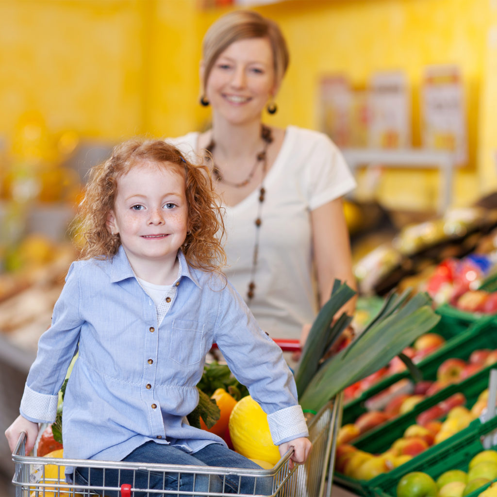 mother in a supermarket pushing a trolley filled with fresh fruit and veg with the daughter riding on the front; 