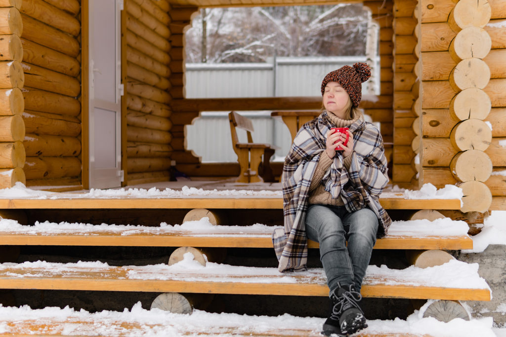 Woman in warm blanket relaxing and drinking morning coffee on the steps log cabin in winter. 
