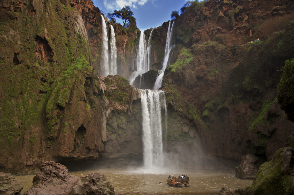 Tourist raft in water dwarfed by 2 tier red rock formation and impressive waterfall