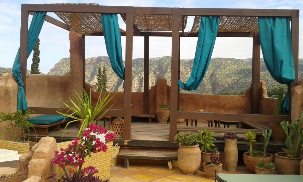 Raised seating area with stone walls, woven roof panels, green curtains and view over rocky hillside