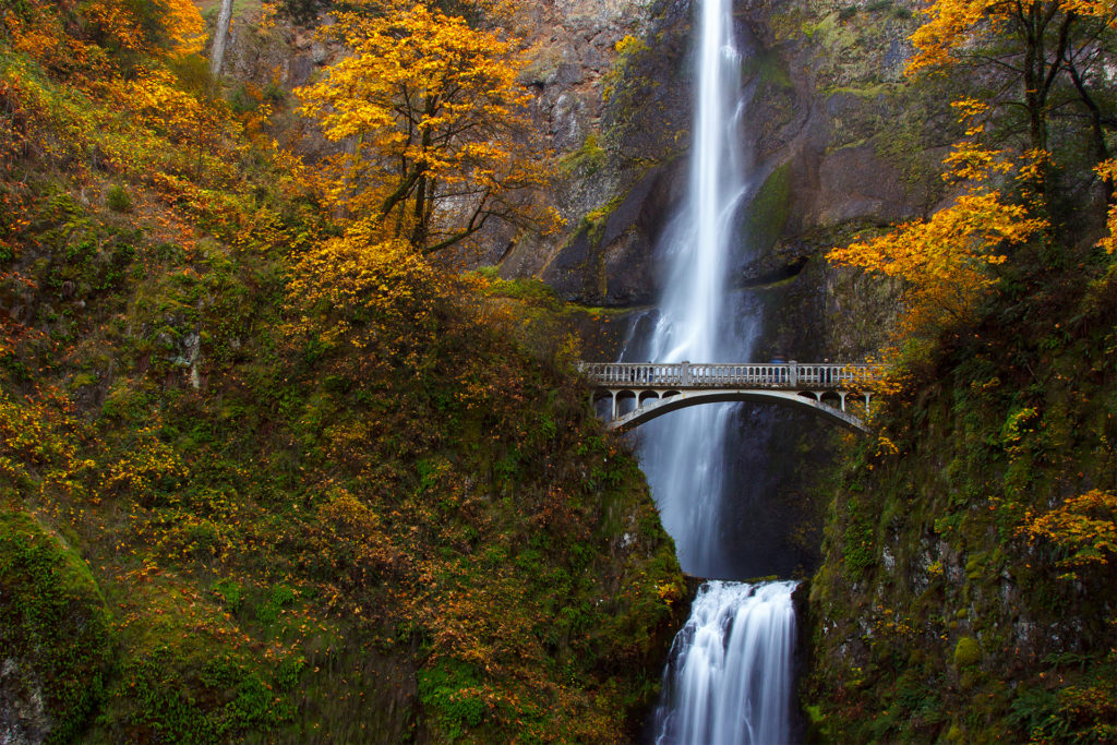 Narrow silvery waterfall plummets hundreds of feet passing metal bridge that crosses in front of it halfway up. Picturesque autumn trees 