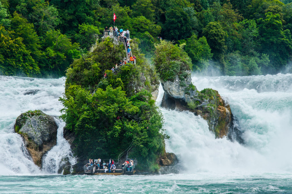 Tall, narrow rocky island amid river and waterfalls. People are climbing steps to the top.