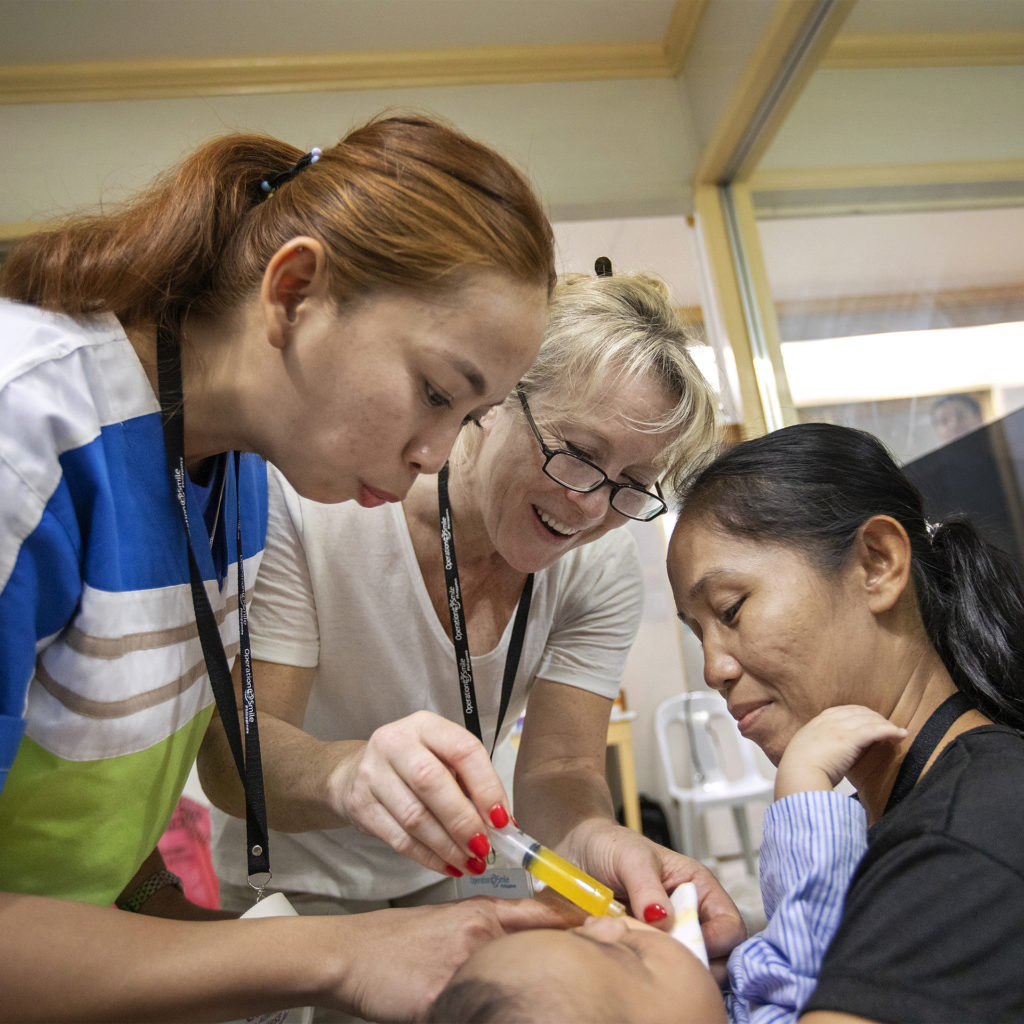 Mother holds baby as nurse and volunteer give medicine in an oral syringe