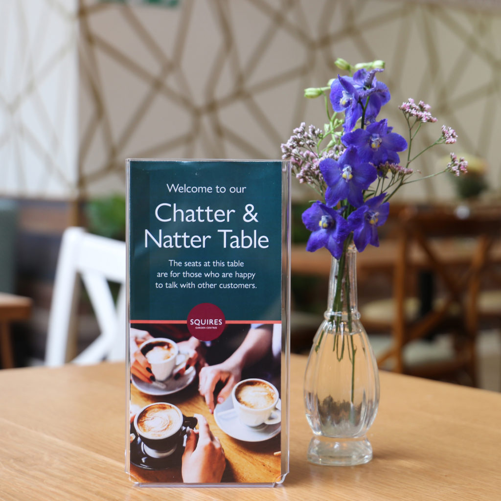 Table in cafe with small vase of flowers and Chatter And Natter sign: This table is for people who are happy to talk to other customers.