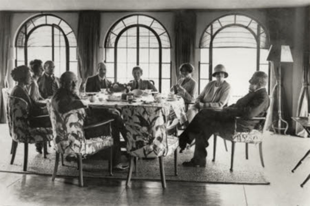 Black and white photo of people sitting around a table in front of 3 arched windows. One woman is wearing a cloche hat.