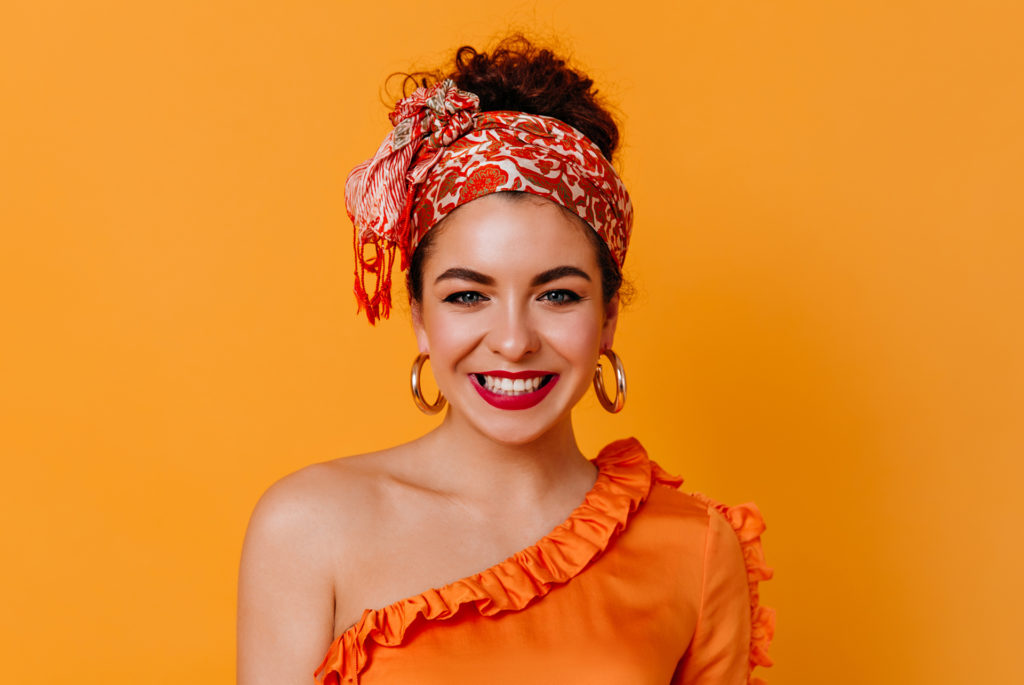 Happy woman with orange patterned silk scarf tied around head, hair visible above, orange background