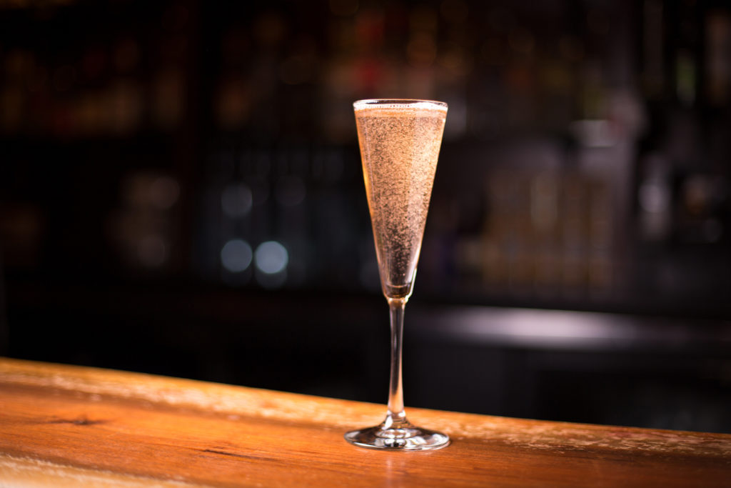 Low angle close up glass of sparkling champagne in traditional crystal flute stemmed wine glass with prosecco bubbles fizzing to surface in dramatic bar light happy hour drink restaurant scene; 