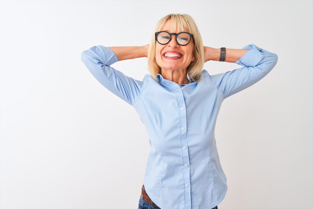 Middle age businesswoman wearing elegant shirt and glasses over isolated white background relaxing and stretching, arms and hands behind head and neck smiling happy