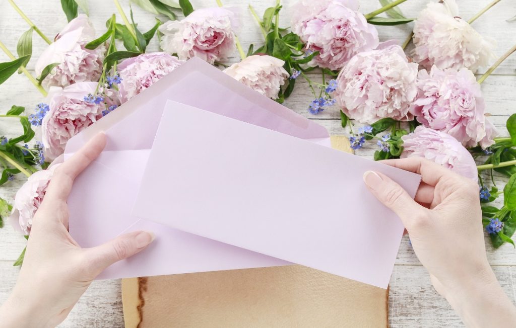 Woman's hands hold pale pink paper and envelope with pale lilac coloured peony flowers on the desk behind