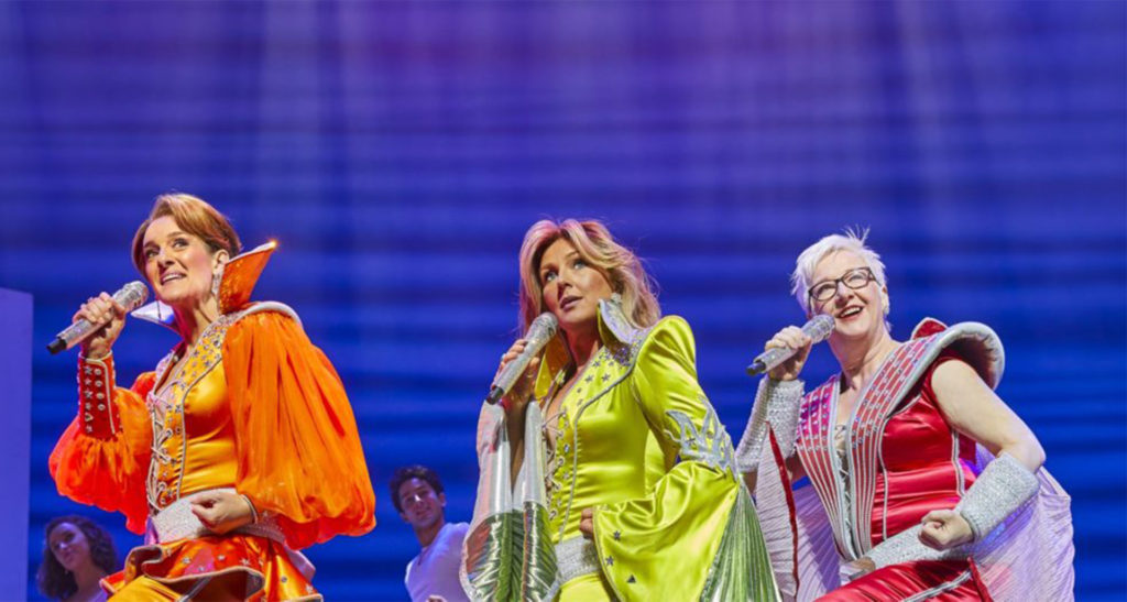 Three cast members of Mamma Mia on stage in the West End in 1970s stage outfits