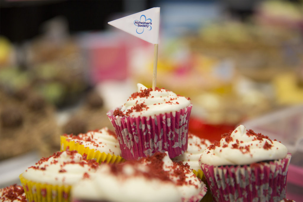 Plate of cupcakes with cream icing and red sprinkles, one with an Alzheimer's Society flag on a cocktail stick. Fundraiser for Alzheimer's Society and Dementia Friends.