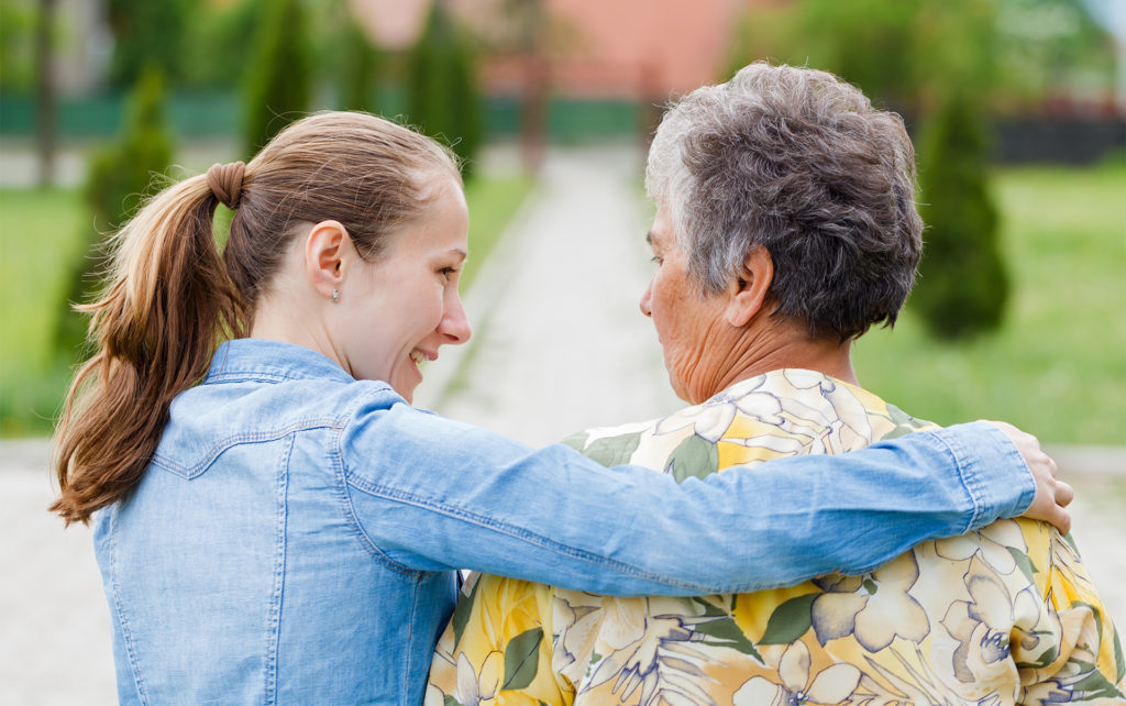 Young woman in denim shirt puts an arm around elderly woman in yellow floral shirt, back view, walking in a garden