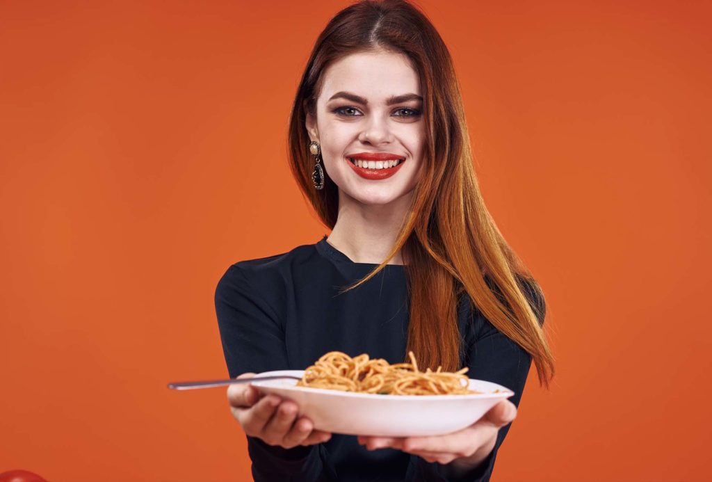A smiling woman is holding a plate with spaghetti and on an orange background.