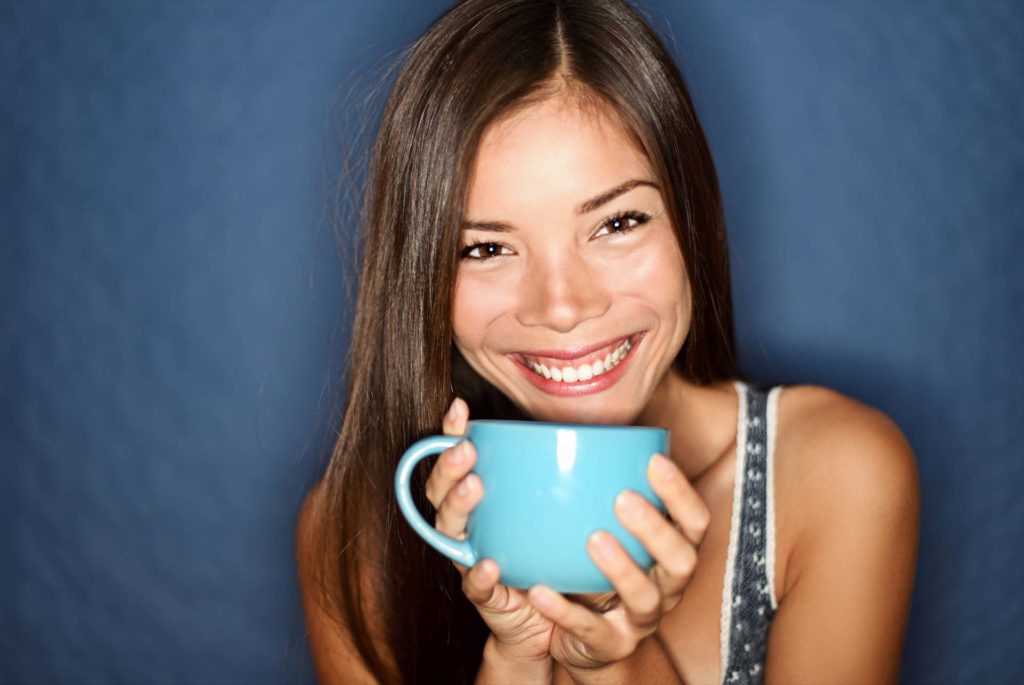 Woman smiling drinking tea on blue background. Young beautiful multiracial Asian / Caucasian female model joyful.