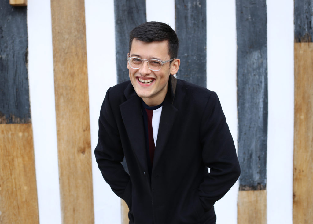 Bake Off contestant Michael Chakraverty, smiling, in a smart black jacket against white, blue and natural planks of wood