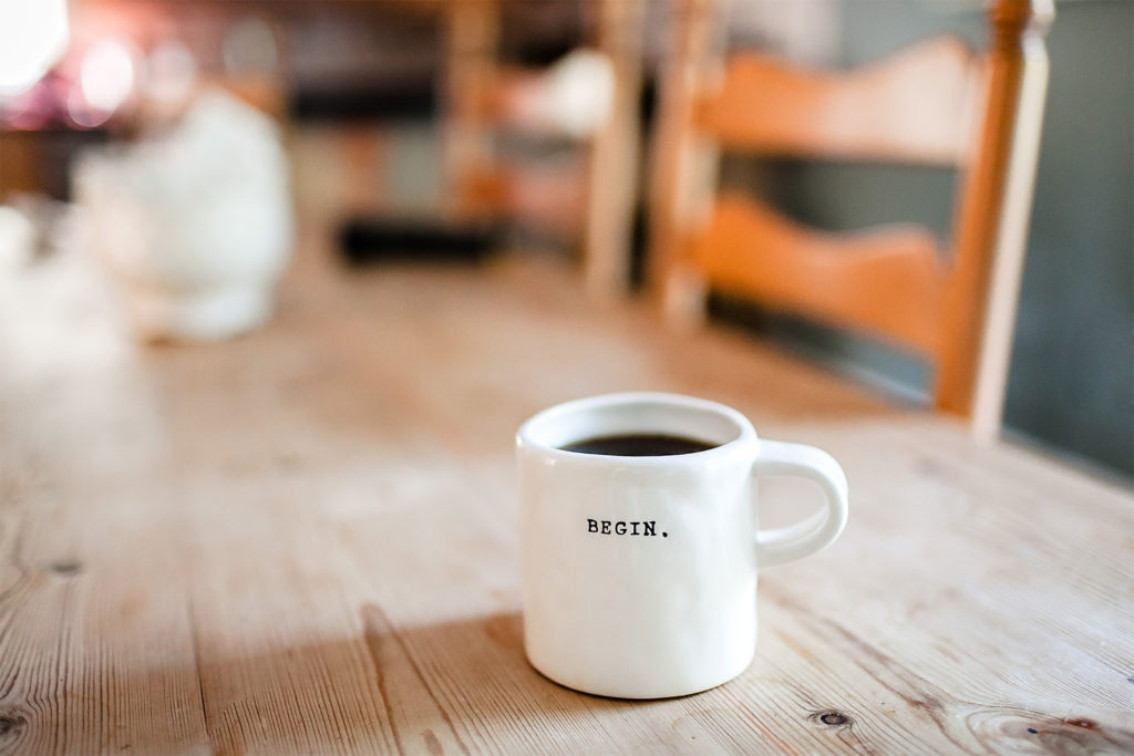 Mug of black coffee on wooden kitchen table. White mug has one word printed in black: Begin. Motivation for new career.