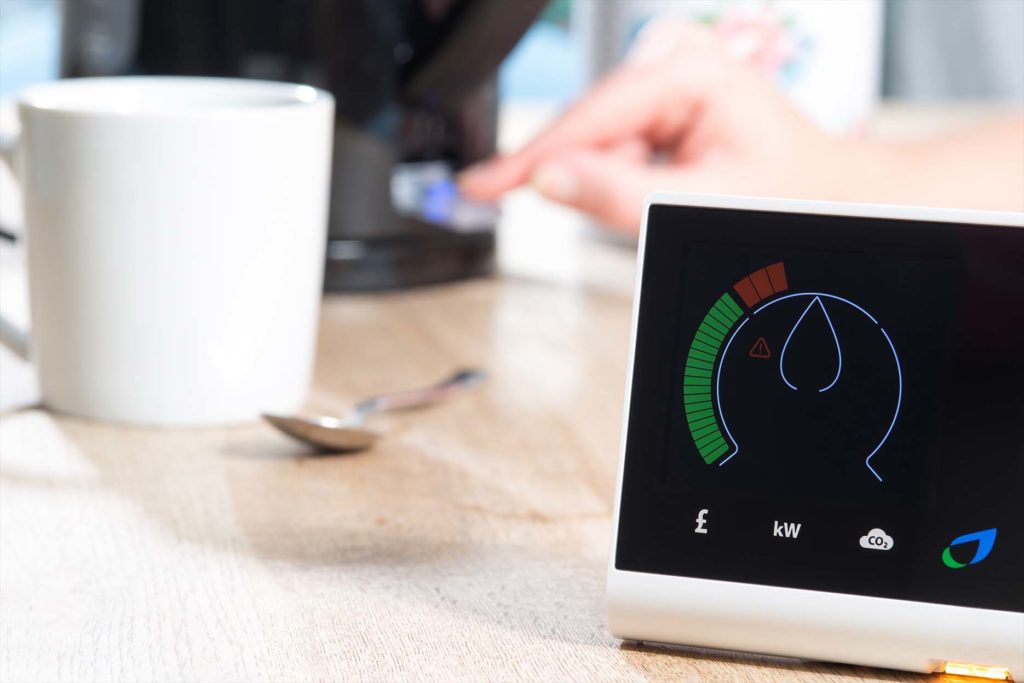 A smart meter is displayed on a wooden surface near mug and spoon and a kettle which is being switched on by a hand. The meter is giving a digital reading of energy consumption.