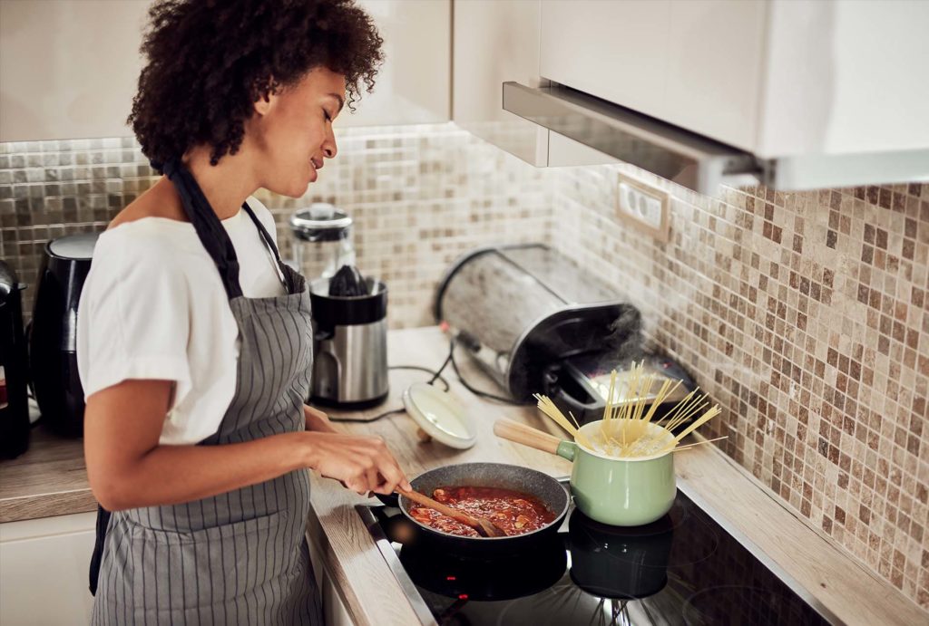 Mixed race woman in apron standing next to stove in domestic kitchen and making pasta and sauce.
