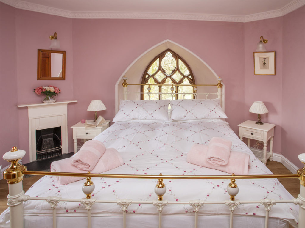 Pink painted bedroom, Gothic pointed window in alcove behind bed head, double bed with brass frame and white covers, small fireplace