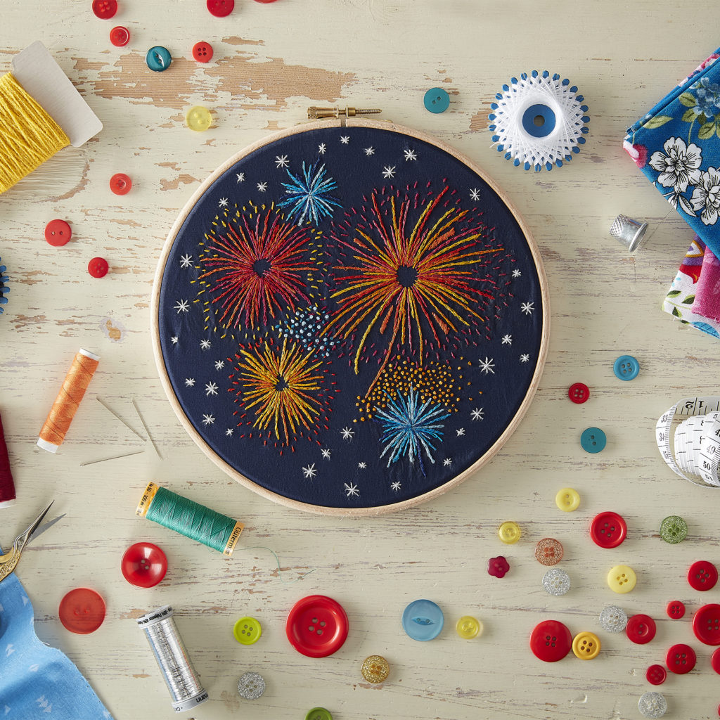 Small circular embroidery frame with black fabric and bright embroidery of fireworks, bright coloured buttons scattered on desk