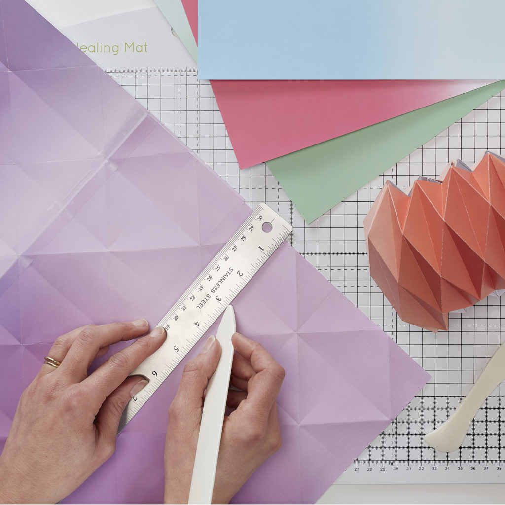 Person with metal ruler and scoring tool, sheets of coloured paper and a piece of honeycomb origami folding