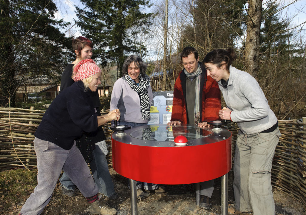 5 people laughing as they turn handles on a red drum shaped gadget, woodland and willow panels behind