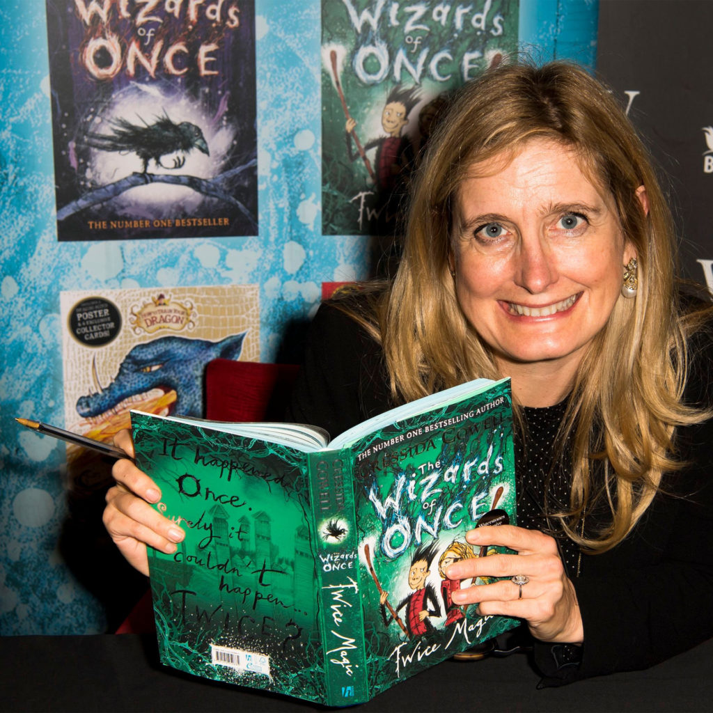 Author Cressida Cowell at an event for her book The Wizards of Once, looking lively, holding book and a pen