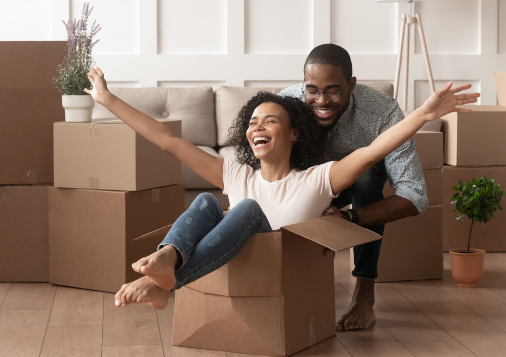 Happy young couple in first home, she is sitting in a packing box and he is pushing it along