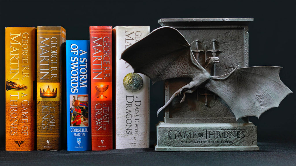 Set of Game Of Thrones books by George Martin standing in a row with special edition DVD set in metal effect box with flying dragon sculpture attached to the front