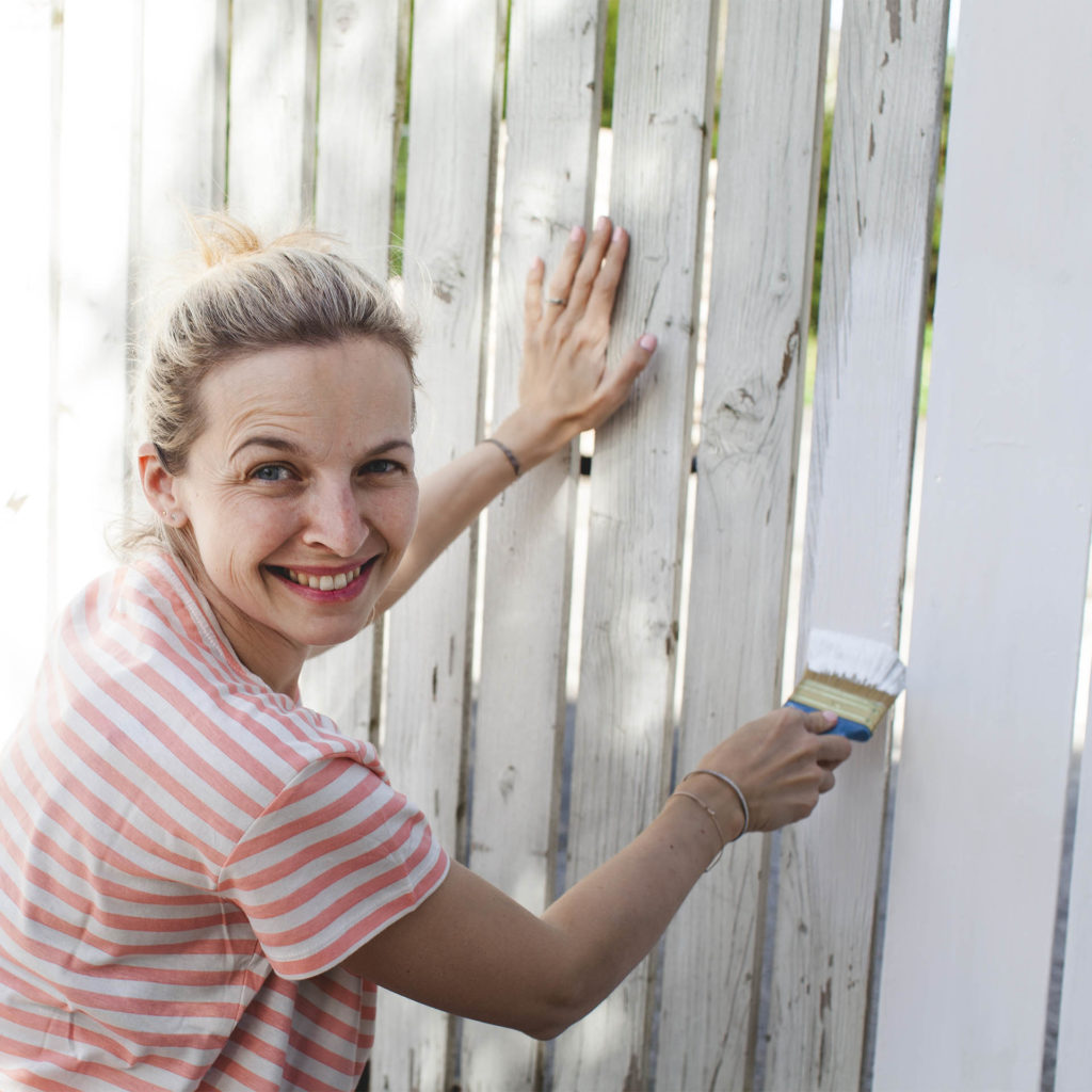 Woman painting wooden fence white on sunny day
