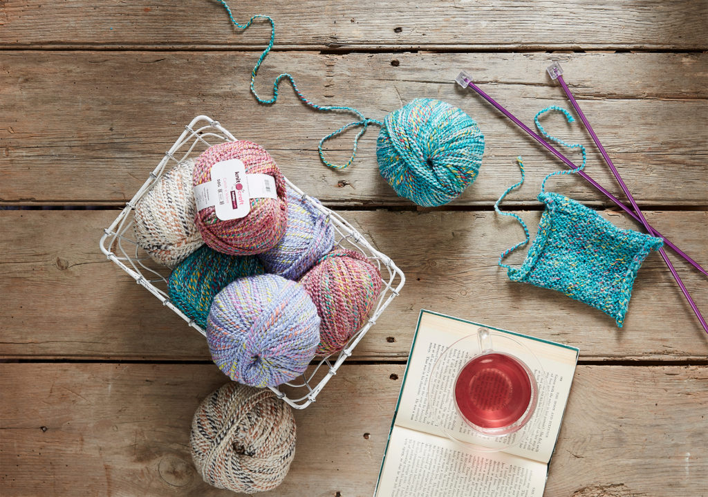 Basket of yarn balls, square knitted in turquoise on knitting needles, also glass of juice and open book