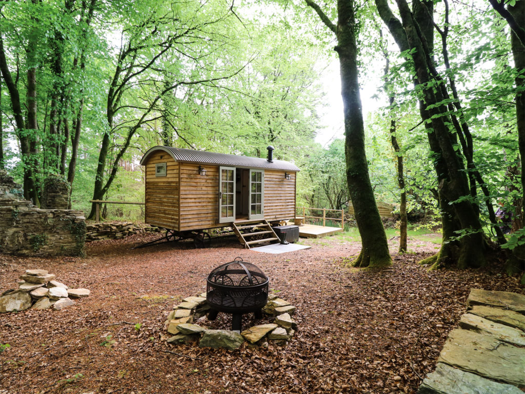 Converted wooden shepherd's hut on wheels in woodland clearing with fire pit