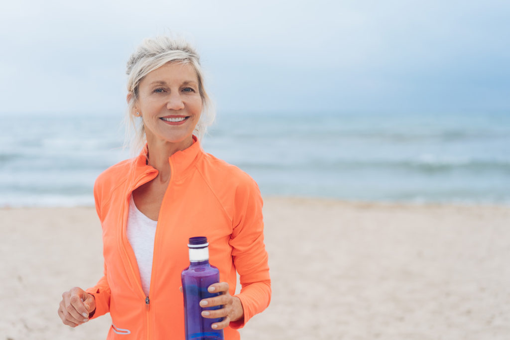 Vivacious fit woman walking on a beach in colorful sportswear carrying a blue bottle of water smiling at the camera; 