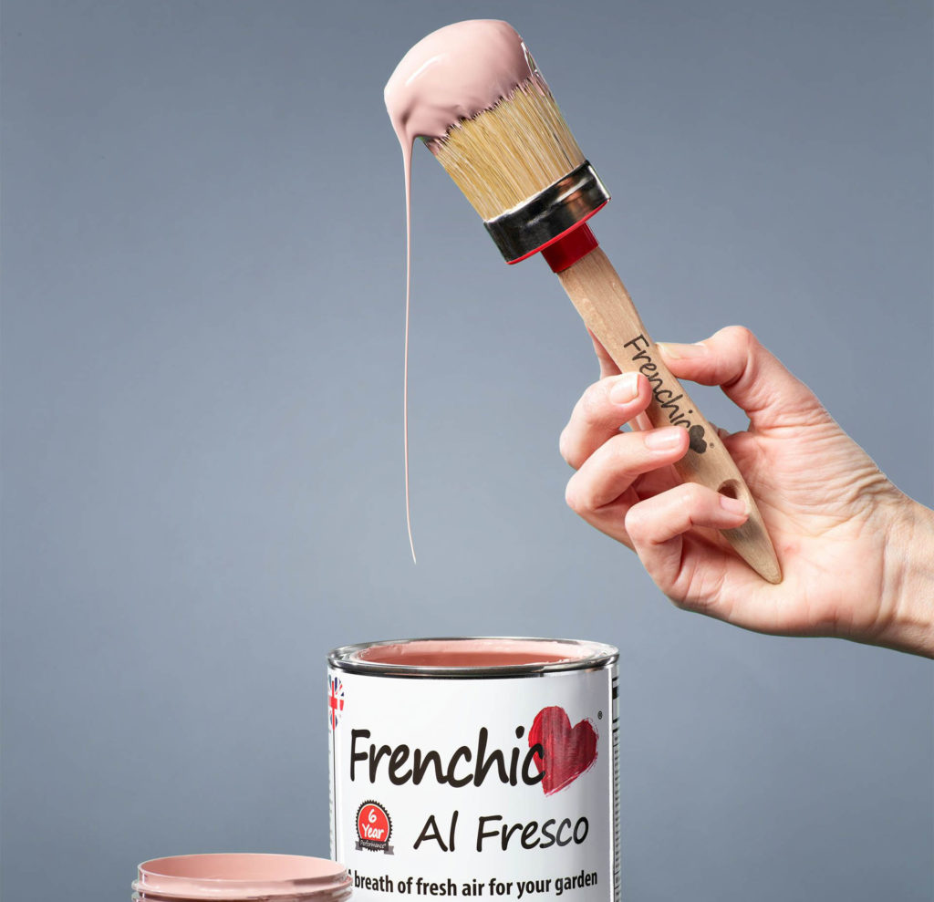 Pot of Frenchic Al Fresco brand paint in Dusky Pink. A woman's hand is holding a paintbrush that has been dipped in the thick, creamy paint.