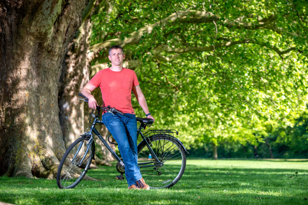 Young man in red t shirt and jeans leans against bicycle under a big tree in the countryside