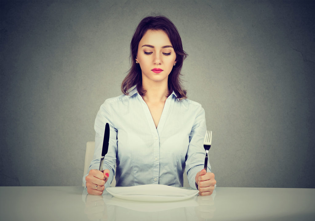Serious woman with fork and knife sitting at table with empty plate isolated on gray wall background