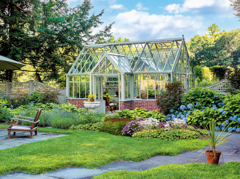 Victorian style greenhouse with several apexes, tall narrow glass panels, white frame, set in beautiful garden