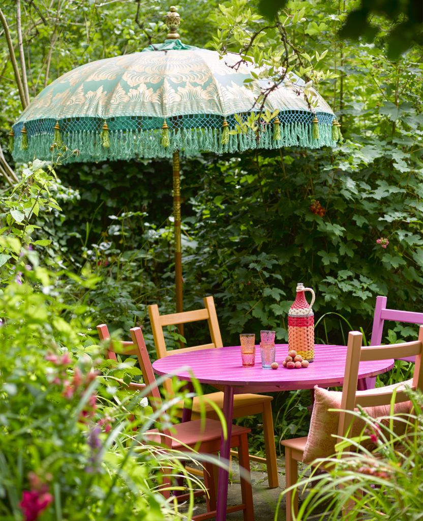 Outdoor table and chairs painted pink and orange with green umbrella and climbing plants all around