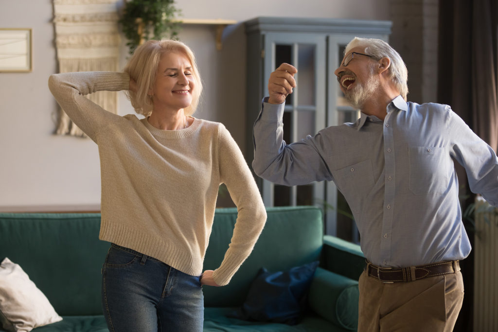 Happy active positive retired elder couple dancing together in living room, cheerful old senior husband and mature middle aged fit wife enjoying fun leisure activity laughing bonding moving at home; 