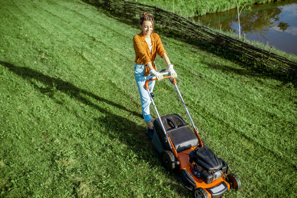 Beautiful young woman cutting grass with gasoline lawn mower, gardening on the backyard in the countryside, view from above; 