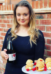 Smiling blonde girl by brick wall outdoors, holding plate of cream puffs and bottle of I Heart Prosecco