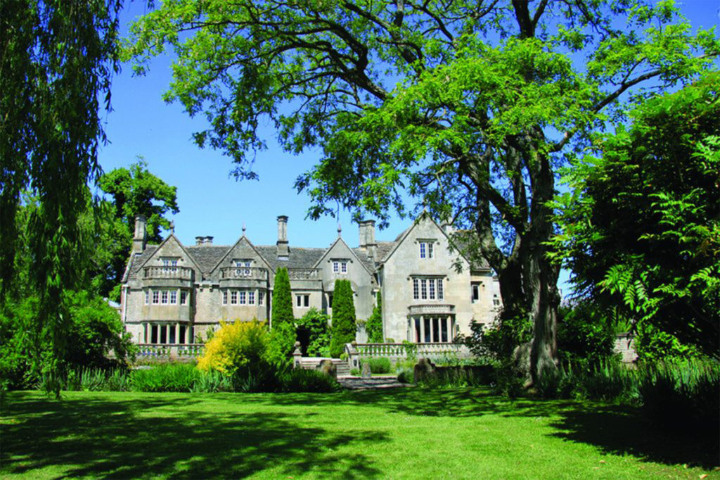 Tradiional grey stone building with mullion windows, seen through lush green trees and gardens
